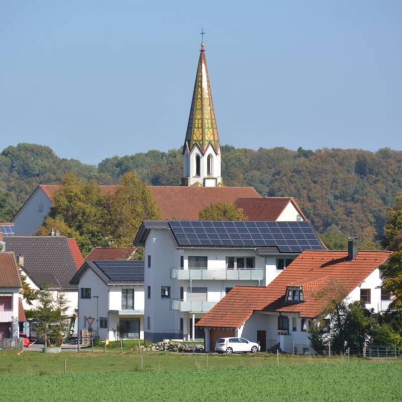 Eine ländliche Ortschaft mit mehreren weißen Häusern und einem Kirchturm im Hintergrund, umgeben von Bäumen und einem blauen Himmel.