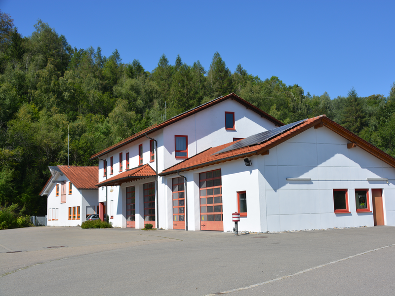 Bauhof und Feuerwehr Warthausen, ein modernes, weißes Gebäude mit roten Dächern steht vor einem bewaldeten Hügel.