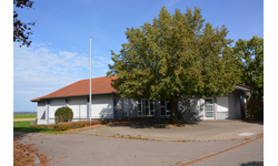 Das Gemeindehaus Oberhöfen, ein flaches, graues Gebäude mit einem roten Ziegeldach steht neben einem großen Baum auf einem asphaltierten Hof.