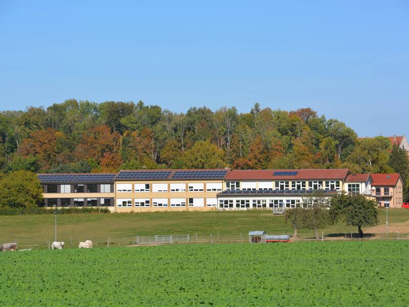 Die Grundschule, ein längliches Gebäude mit Solarpanelen auf dem Dach steht vor einem Wald, umgeben von grünen Feldern und vereinzelten Tieren auf der Wiese.