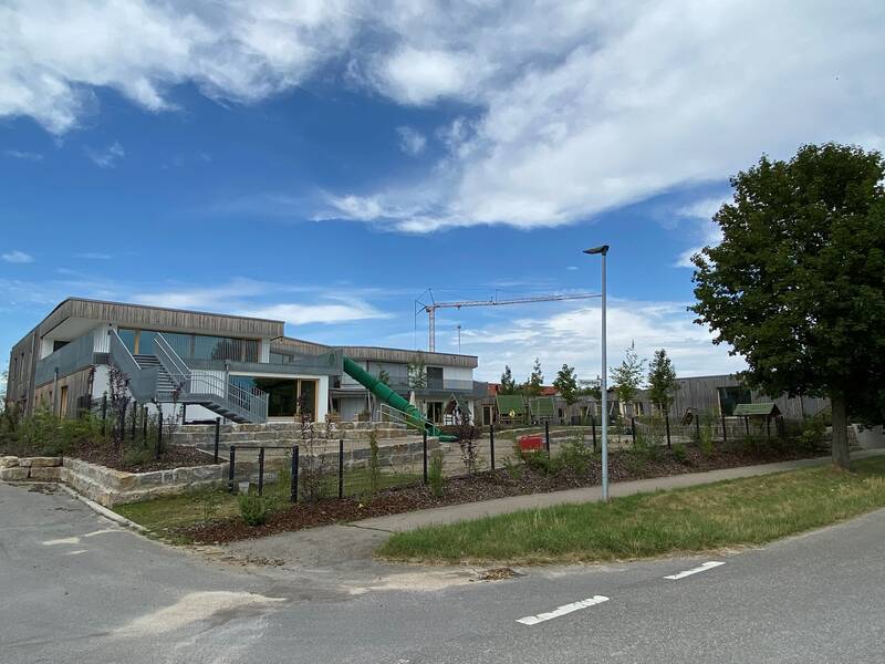 Das Bild zeigt die moderne Kindertagestätte Birkenhard mit Außentreppen, einem Spielplatz mit Rutsche und einem Kran im Hintergrund unter einem teils bewölkten Himmel.