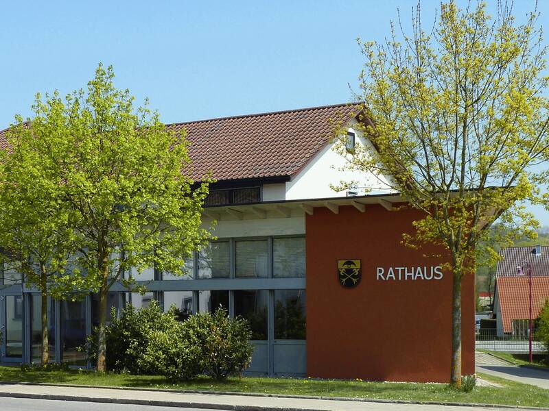 Das Bild zeigt ein modernes Rathausgebäude mit roten Ziegeldächern, umgeben von Bäumen und einem Bushaltestellenschild im Vordergrund.