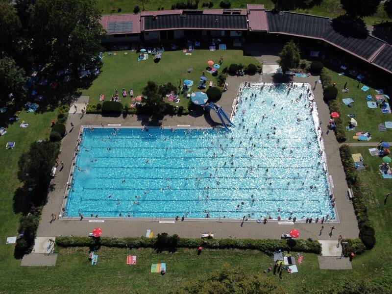 Luftbild des großen, belebten Freibads Warthausens mit vielen schwimmenden und sich entspannenden Besuchern auf umgebenden Liegewiesen.