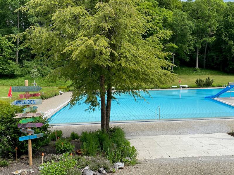 Das große Schwimmbecken mit einer Rutsche des Freibads Warthausens befindet sich in einer grünen, von Bäumen umgebenen Parkanlage, neben einem Holzwegweiser.