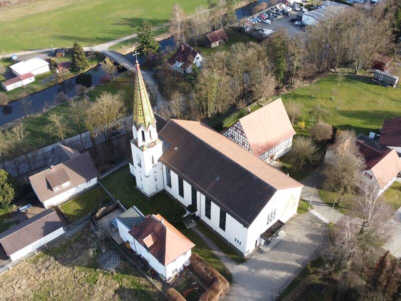 Eine Luftaufnahme der katholischen Kirche Warthausens mit einem hohen Turm und umliegenden Gebäuden sowie teilweise bewaldeter Landschaft im Hintergrund.