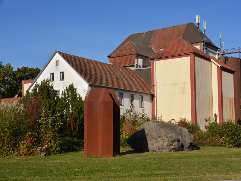 Eine Mischung aus traditionellen und industriellen Gebäuden steht in einer grünen, bewachsenen Landschaft mit einer großen Metallskulptur und einem Fels im Vordergrund.