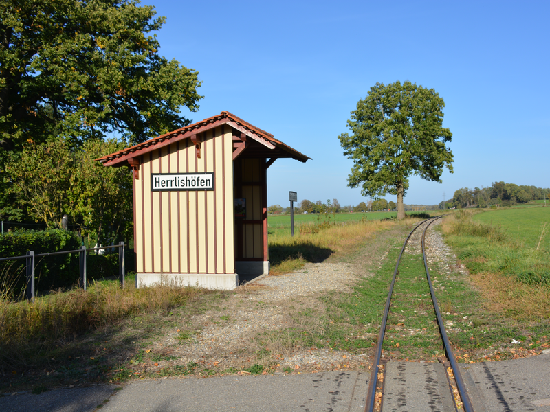 Ein kleines Gebäude mit der Aufschrift "Herrlishöfen" steht neben einer schmalen Eisenbahnschiene, umgeben von Natur und einem einzelnen Baum.