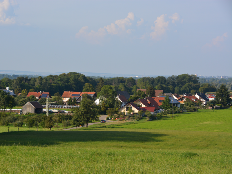 Eine ländliche Szene mit einem kleinen Dorf, umgeben von grünen Wiesen und Bäumen, unter einem blauen Himmel mit einigen Wolken.