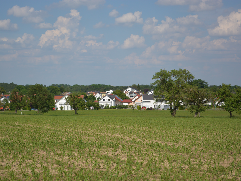 Ein kleines Dorf mit mehreren weißen Häusern und roten Dächern liegt eingebettet in einer grünen Landschaft aus Feldern und Bäumen unter einem blauen Himmel mit zahlreichen Wolken.