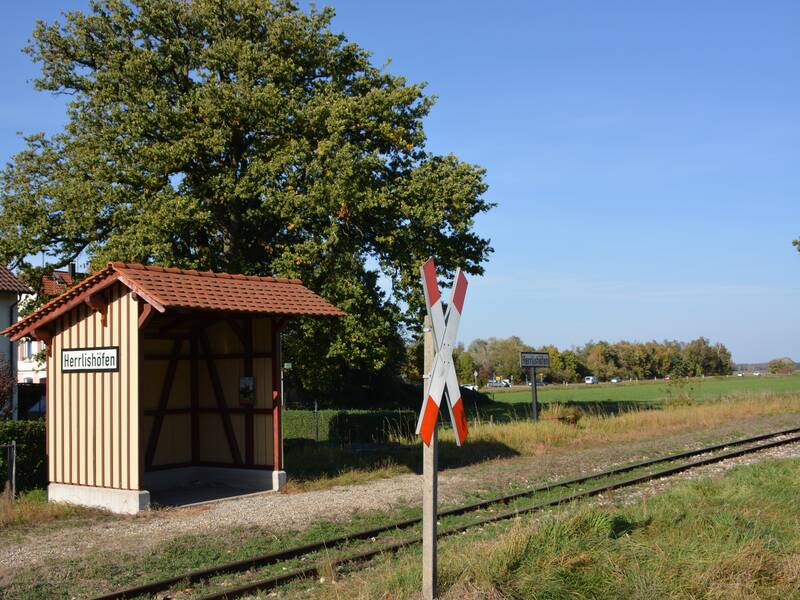 Das Bild zeigt einen kleinen überdachten Wartebereich mit der Aufschrift "Herrlishöfen" neben Bahngleisen und einem Andreaskreuz, umgeben von grüner Landschaft und Bäumen.