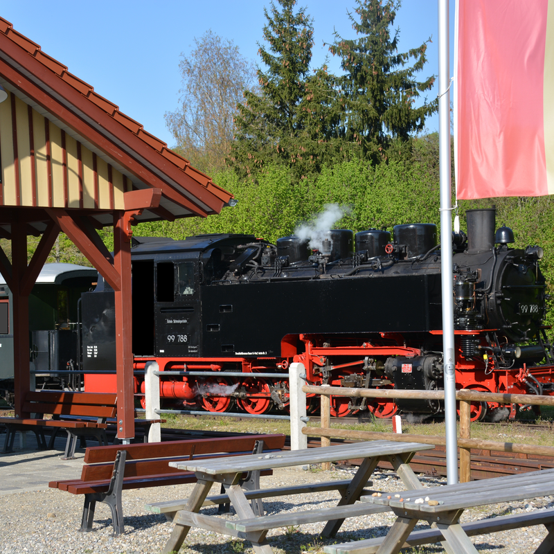 Eine schwarze Dampflokomotive steht am Bahnhof Warthausen neben einem Holzhäuschen und einer rot-gelben Flagge, während im Vordergrund Bänke und Tische zu sehen sind.