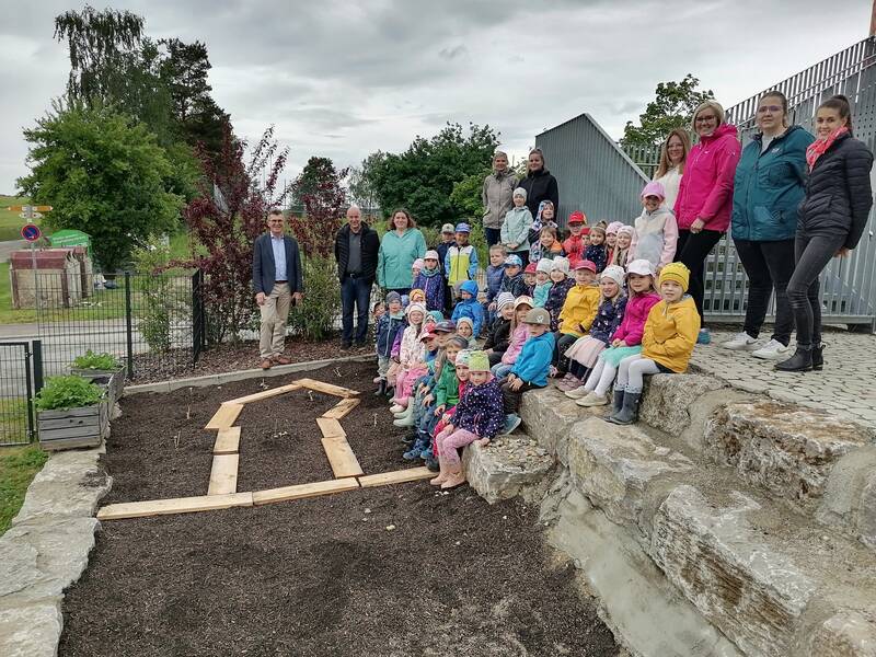 Eine Gruppe von Kindern und Erwachsenen steht und sitzt in einem Garten neben einem mit Holzlatten abgesteckten Beet.