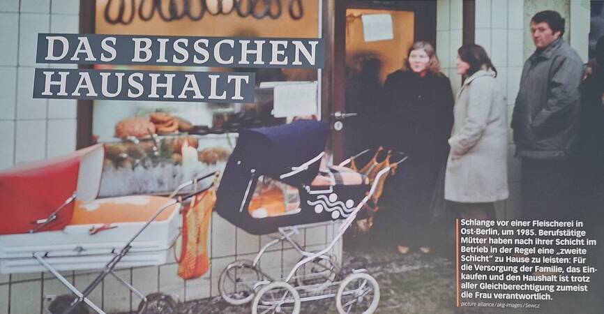 Vor einem Laden mit der Aufschrift "Das bisschen Haushalt" stehen mehrere Personen in einer Schlange, und im Vordergrund sind zwei Kinderwagen zu sehen.
