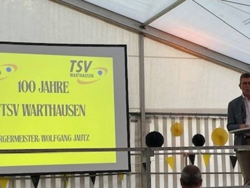 Ein Mann hält eine Rede an einem Rednerpult neben einer gelben Leinwand, die das 100-jährige Jubiläum des TSV Warthausen ankündigt.