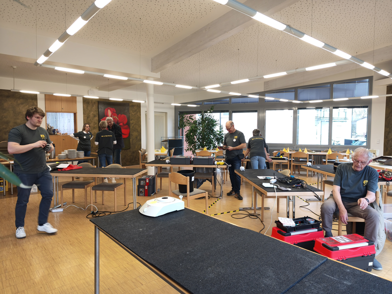 Mehrere Personen arbeiten in einem hellen Raum, der mit Tischen und Stühlen ausgestattet ist, und sie scheinen Geräte zu reparieren oder zu warten.