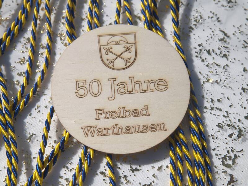 Ein rundes Holzschild mit der Gravur "50 Jahre Freibad Warthausen" liegt auf glitzerndem Hintergrund, umgeben von blau-gelb geflochtenen Schnüren.