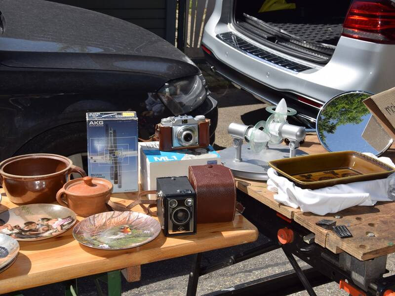 Ein Flohmarktstand zeigt verschiedene antike Gegenstände, darunter Kameras, Porzellanteller und Töpferwaren, vor geparkten Autos.