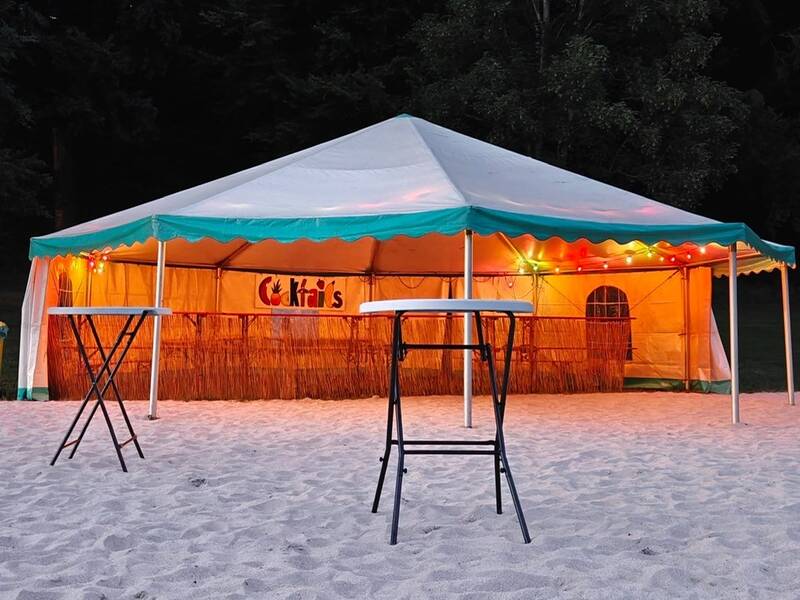 Ein festlich beleuchtetes Zelt auf einem Sandboden ist mit einer Bar und zwei Stehtischen davor aufgebaut.