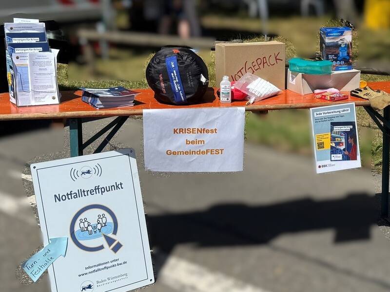 Ein Informationsstand auf einem GemeindeFest zeigt Flyer und Notfallausrüstung, inklusive eines Notfalltreffpunktschildes.