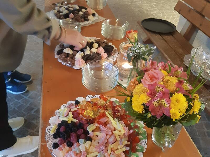 Ein Tisch mit verschiedenen Schalen voller Süßigkeiten und Blumen, daneben Kerzen und ein Mensch, der nach Süßigkeiten greift.