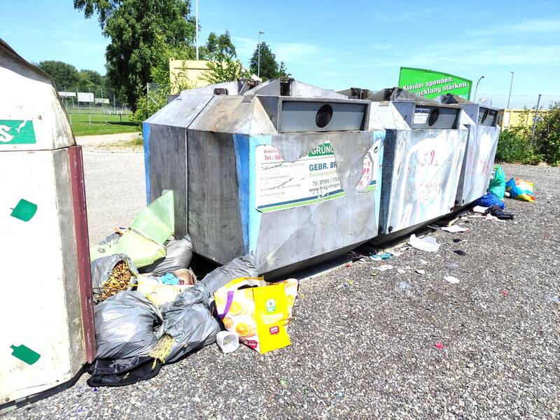 Neben großen Altglascontainern liegt eine Ansammlung von Müllsäcken und losem Abfall auf dem Boden.
