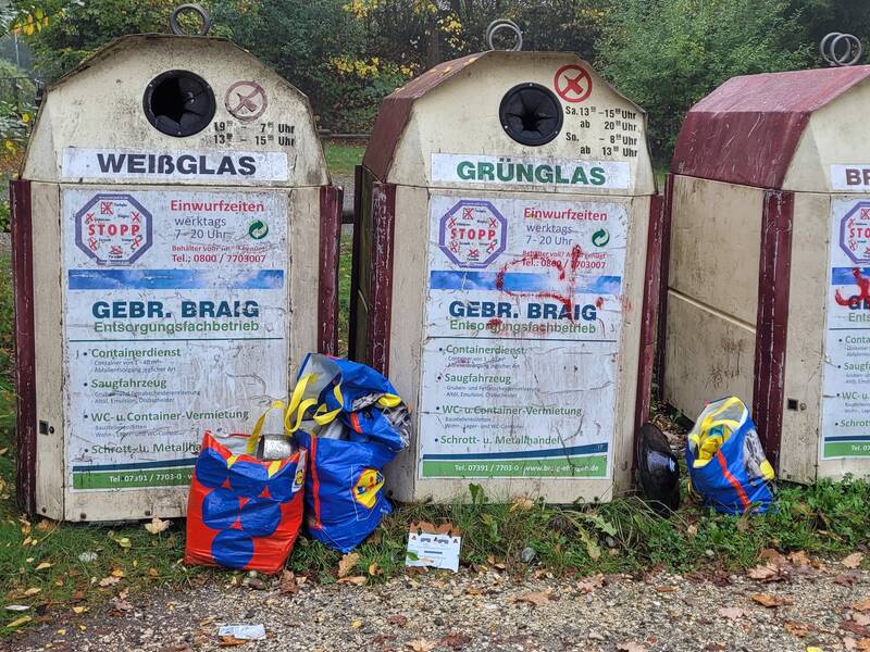 Drei Altglascontainer für Weiß-, Grün- und Braunglas stehen nebeneinander, umgeben von bunten Taschen, vor einem Hintergrund aus Herbstbäumen.