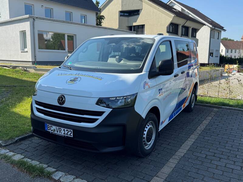 Ein weißer VW-Transporter mit Aufschriften steht auf einem gepflasterten Parkplatz vor mehreren Gebäuden.