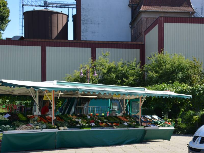 Ein Marktstand mit frischem Obst und Gemüse steht vor einer Hintergrundkulisse von Industriegebäuden und umgeben von grüner Vegetation; rechts ist ein weißer Lieferwagen zu sehen.
