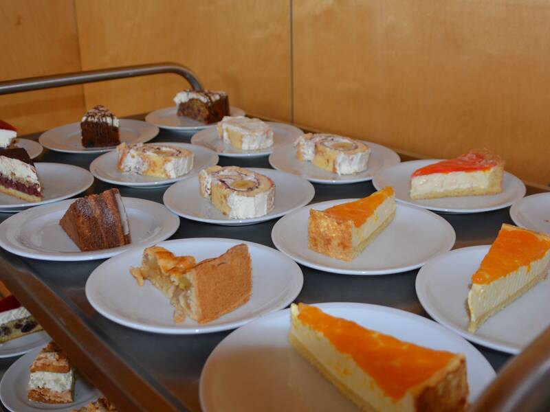 Auf einem Tisch oder Wagen stehen mehrere Teller mit verschiedenen Kuchenstücken, darunter Obstkuchen, Sahnetorten und Schokoladenkuchen.