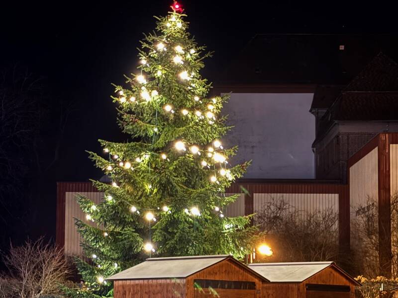 Ein großer, festlich mit Lichtern geschmückter Weihnachtsbaum steht im Dunkeln neben zwei hölzernen Verkaufshütten auf einem gepflasterten Platz.
